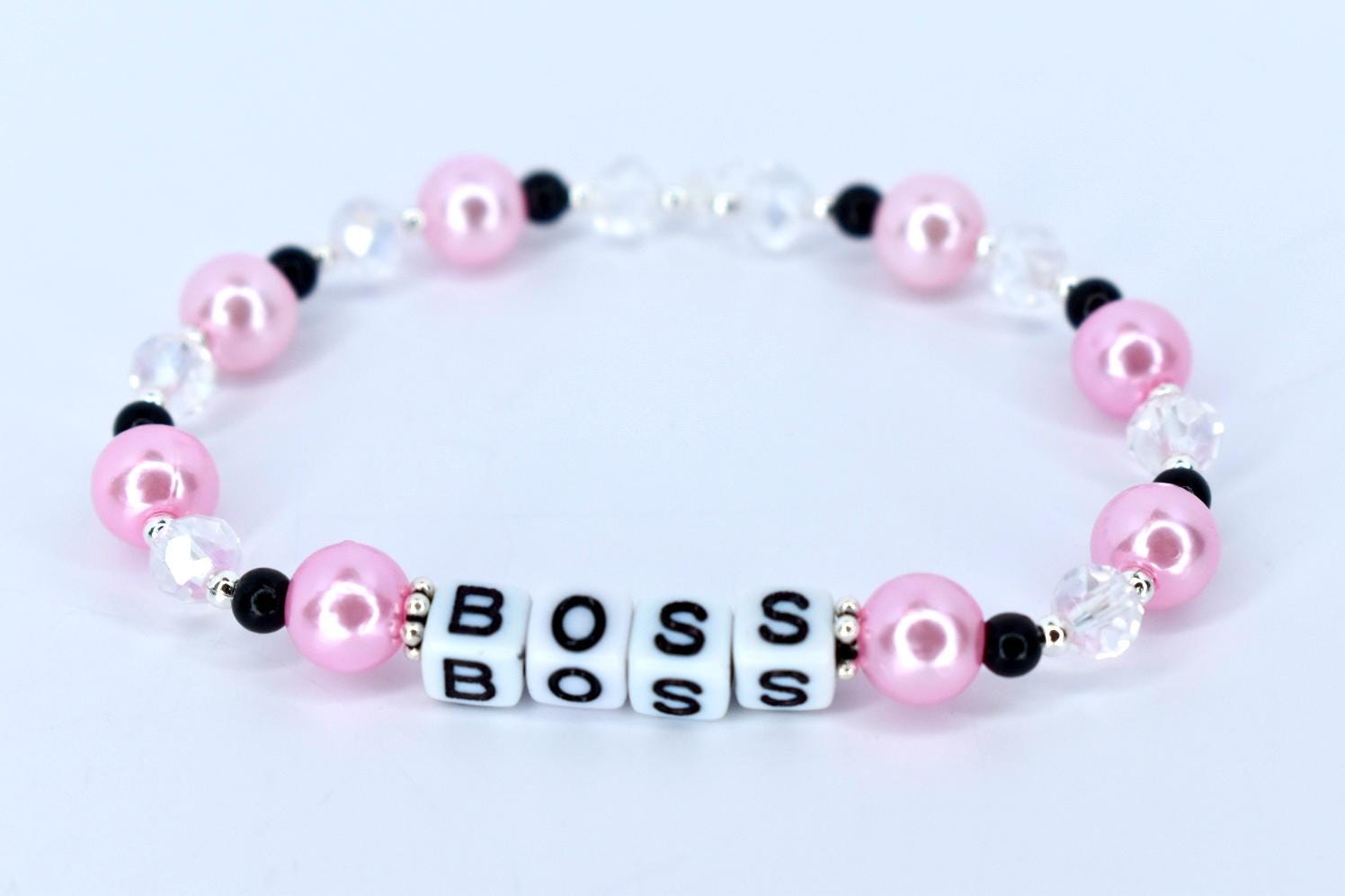 A pink and white beaded bracelet with the word 'BOSS' spelled out in white letter beads. The bracelet features pink pearl beads, clear crystal beads, and black accent beads. The beads are strung together on a stretchy cord, forming a circular shape.