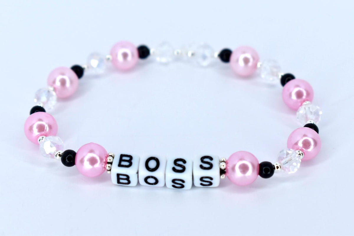 A pink and white beaded bracelet with the word 'BOSS' spelled out in white letter beads. The bracelet features pink pearl beads, clear crystal beads, and black accent beads. The beads are strung together on a stretchy cord, forming a circular shape.