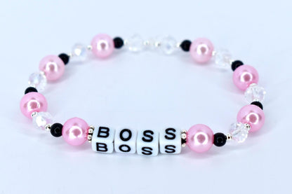 A pink and white beaded bracelet with the word 'BOSS' spelled out in white letter beads. The bracelet features pink pearl beads, clear crystal beads, and black accent beads. The beads are strung together on a stretchy cord, forming a circular shape.