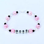 A pink beaded bracelet with the word 'BOSS' spelled out in white letter beads. The bracelet features pink, black, and clear beads. The pink beads are round and the clear beads are faceted. The bracelet is on a white background.