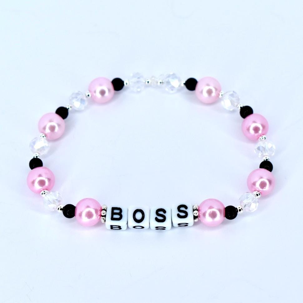A pink beaded bracelet with the word 'BOSS' spelled out in white letter beads. The bracelet features pink, black, and clear beads. The pink beads are round and the clear beads are faceted. The bracelet is on a white background.