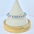 A beaded bracelet with the word "TEACHER" spelled out in white letter beads. The bracelet features colorful beads in shades of blue, pink, yellow, and red. The bracelet is displayed on a light-colored, cone-shaped stand.