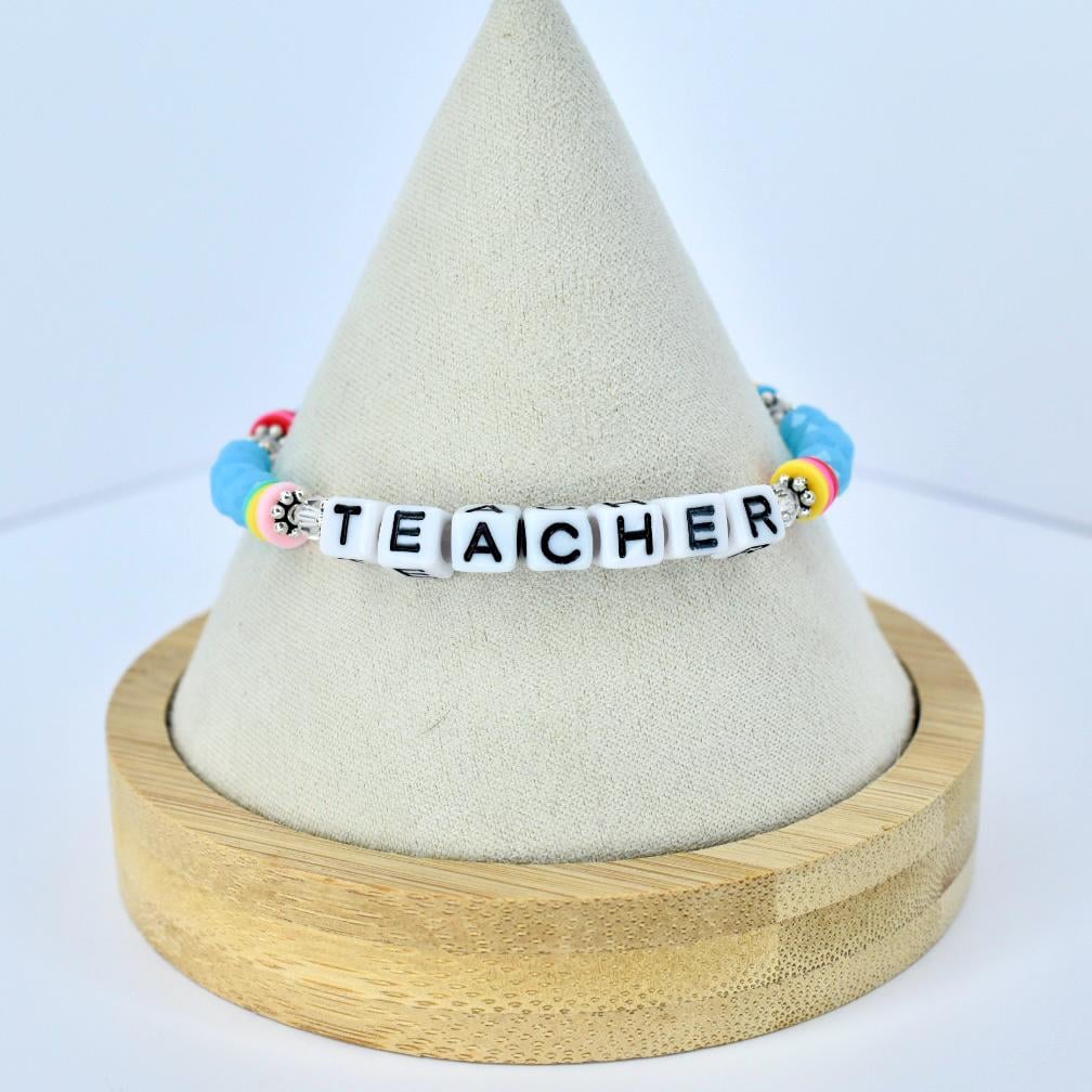 A beaded bracelet with the word "TEACHER" spelled out in white letter beads. The bracelet features colorful beads in shades of blue, pink, yellow, and red. The bracelet is displayed on a light-colored, cone-shaped stand.