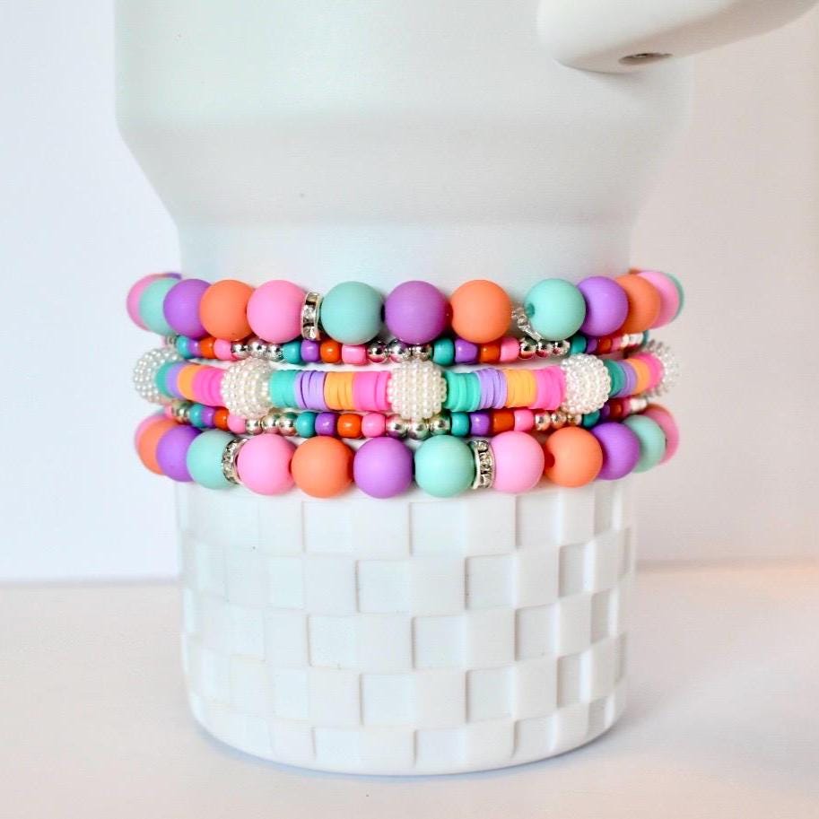 Stack of colorful beaded bracelets in shades of pink, orange, purple, and turquoise. The bracelets feature a mix of round beads and flat disc beads, with silver accents and white pearl-like embellishments. The bracelets are stacked on a white textured surface.