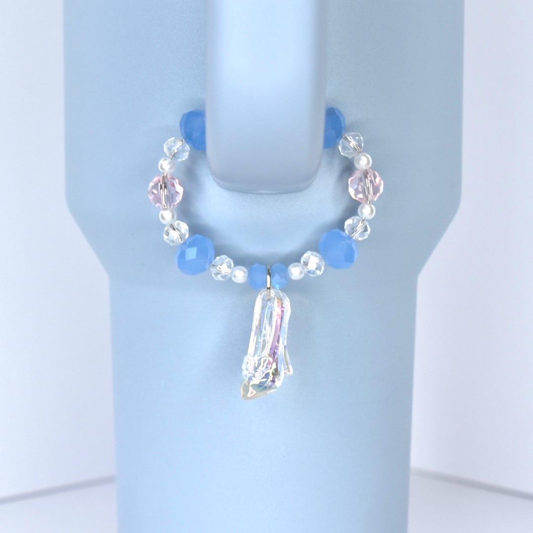 A blue and pink beaded charm bracelet with a clear, iridescent slipper pendant. The bracelet features blue, pink, and clear beads. The charm is attached to a light blue object.