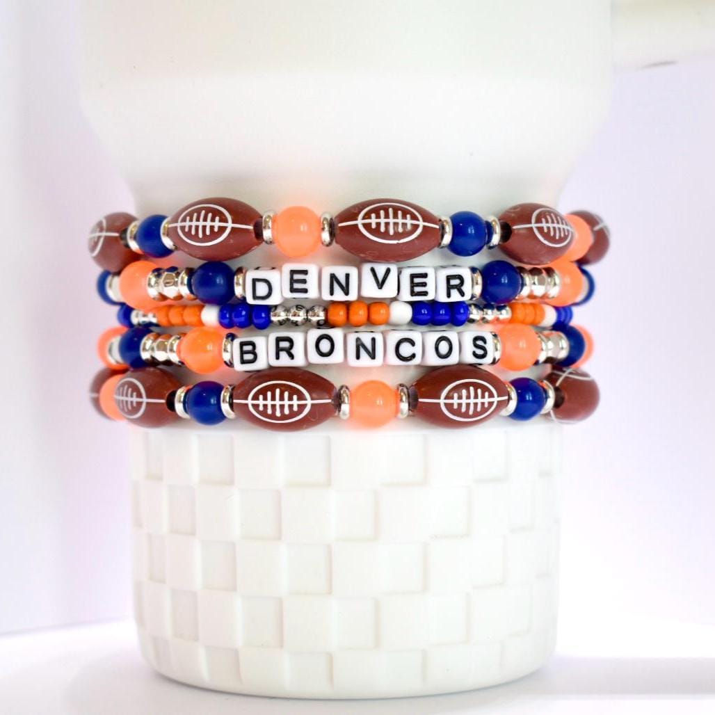 Four stacked beaded bracelets featuring the words 'DENVER BRONCOS'. The bracelets are made with white letter beads, football-shaped beads, orange beads, and blue beads. The bracelets are displayed on a white, textured surface.