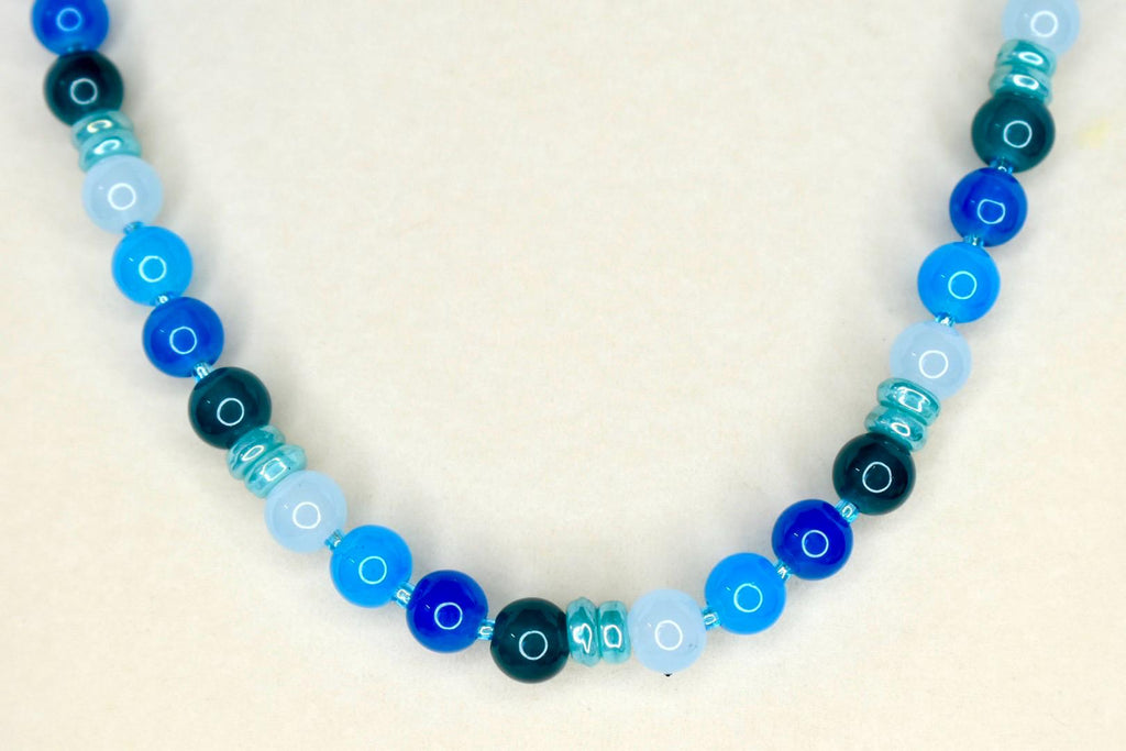 A beaded necklace featuring a gradient of blue hues. The necklace showcases a variety of round beads in shades of light blue, medium blue, and dark blue, interspersed with smaller, textured beads. The necklace is a jewelry item.