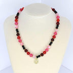 A beaded necklace with a gradient of colors, including red, pink, and black beads. The necklace has a gold-colored pendant with a smiley face design. The necklace is displayed on a white mannequin.