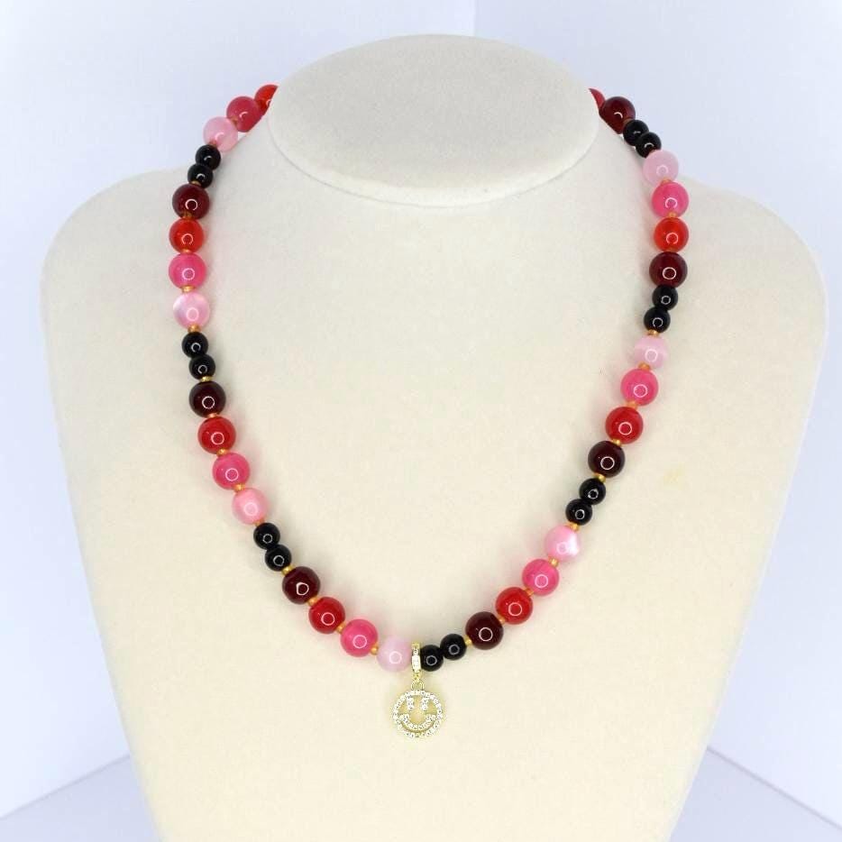 A beaded necklace with a gradient of colors, including red, pink, and black beads. The necklace has a gold-colored pendant with a smiley face design. The necklace is displayed on a white mannequin.