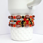 A stack of beaded bracelets featuring football-shaped beads and letter beads spelling 'SAN FRANCISCO 49ERS'. The bracelets are in red, black, and gold colors. The football beads have white stitching details. The bracelets are stacked on a white, textured surface.