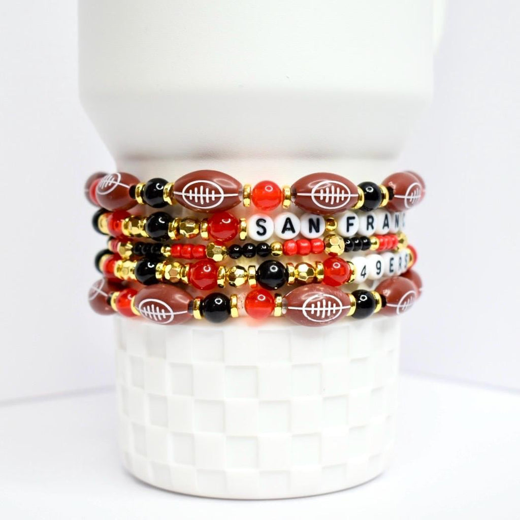 A stack of beaded bracelets with a football theme. The bracelets feature brown football-shaped beads, red, black, and gold beads. One bracelet spells out 'SAN FRAN 49ERS' with white letter beads. The bracelets are stacked on a white, textured surface.