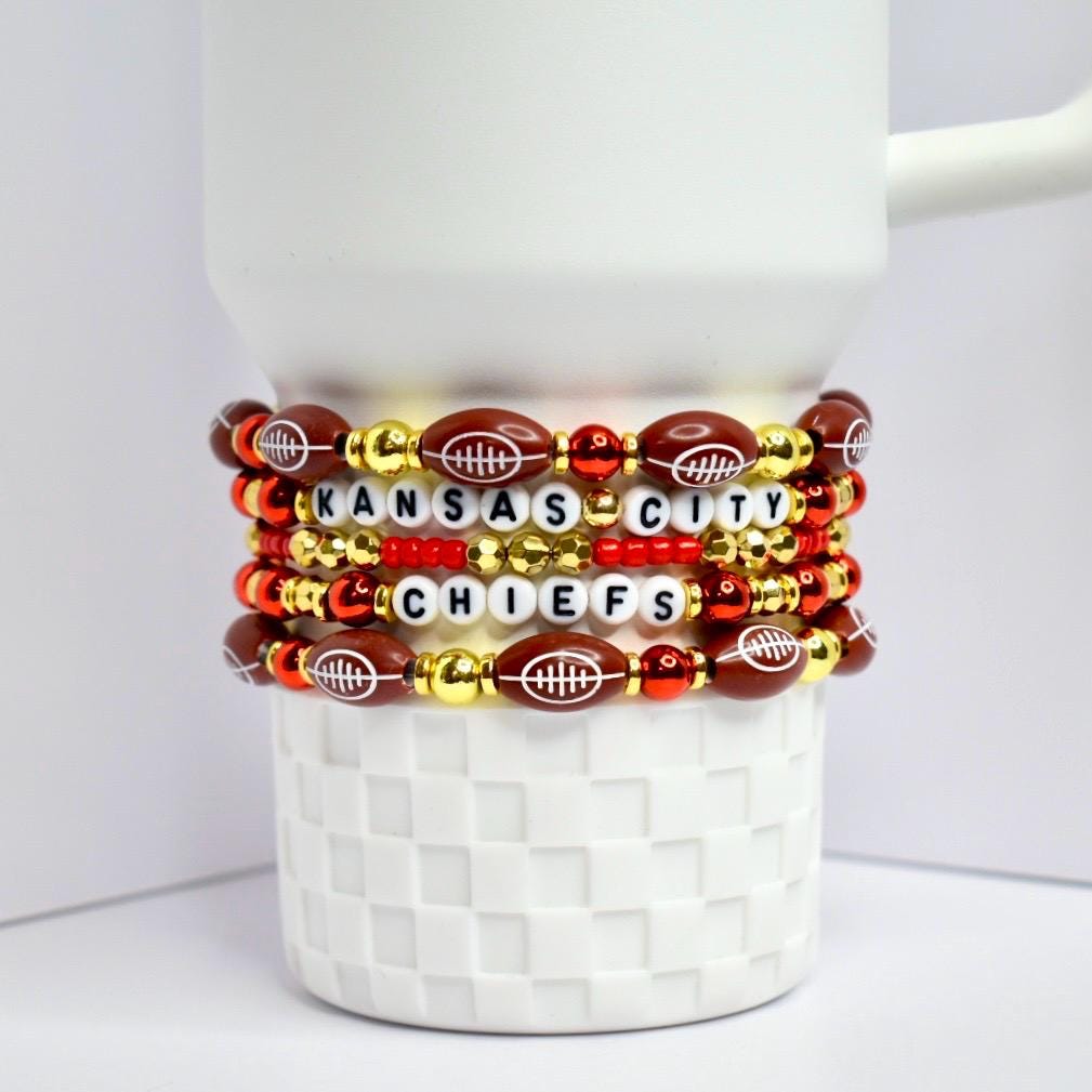 A stack of beaded bracelets featuring the Kansas City Chiefs. The bracelets are arranged on a white, textured surface. The bracelets incorporate football-shaped beads, gold beads, red beads, and white letter beads spelling out 'KANSAS CITY CHIEFS.'