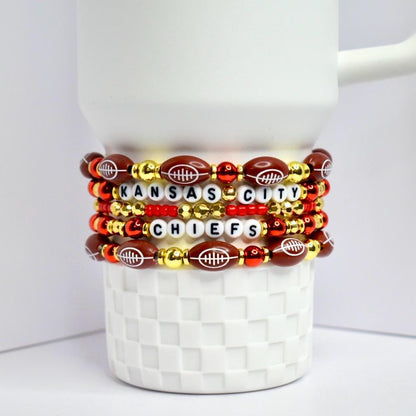 A stack of beaded bracelets featuring the Kansas City Chiefs. The bracelets are arranged on a white, textured surface. The bracelets incorporate football-shaped beads, gold beads, red beads, and white letter beads spelling out 'KANSAS CITY CHIEFS.'
