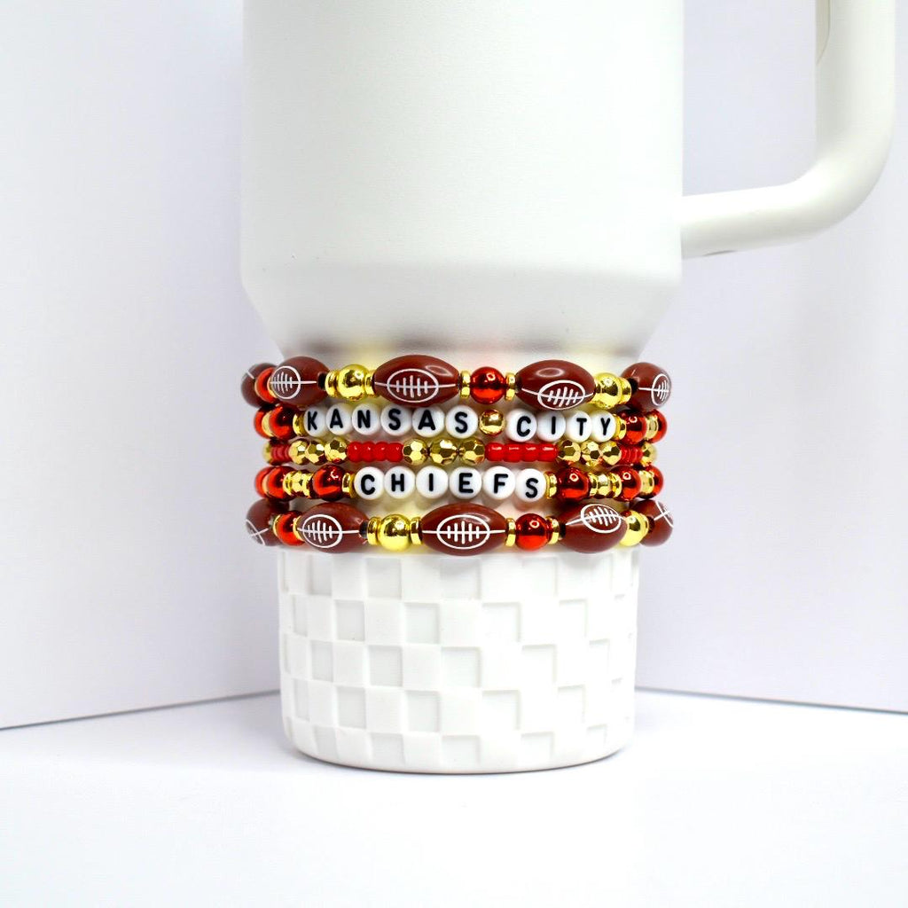 Stack of beaded bracelets wrapped around a white mug. The bracelets feature football-shaped beads, red and gold beads, and letter beads spelling 'KANSAS CITY CHIEFS'. The bracelets are red, gold, and brown with white lettering.