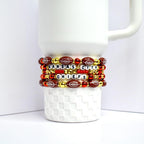 Stack of beaded bracelets wrapped around a white mug. The bracelets feature football-shaped beads, red and gold beads, and letter beads spelling 'KANSAS CITY CHIEFS'. The bracelets are red, gold, and brown with white lettering.