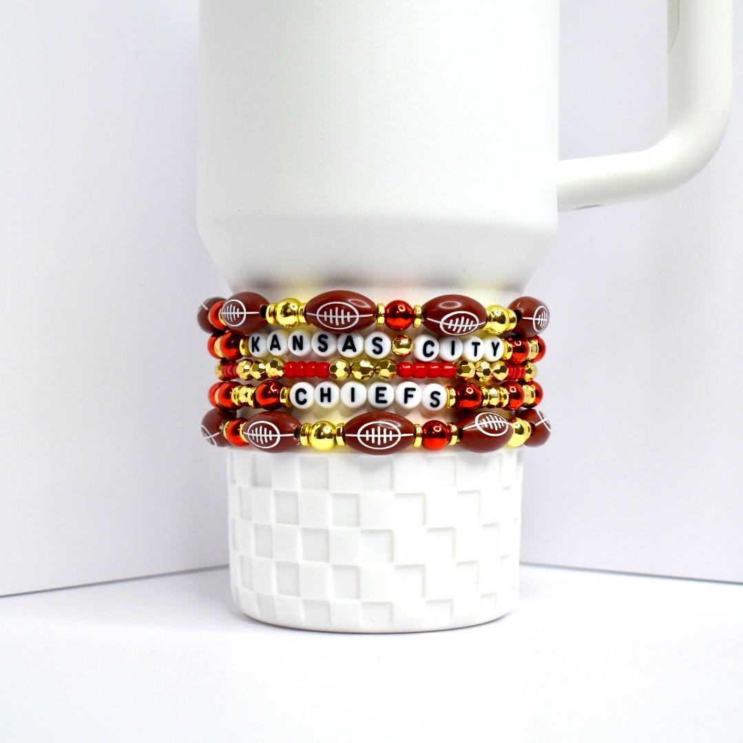 Stack of beaded bracelets wrapped around a white mug. The bracelets feature football-shaped beads, red and gold beads, and letter beads spelling 'KANSAS CITY CHIEFS'. The bracelets are red, gold, and brown with white lettering.
