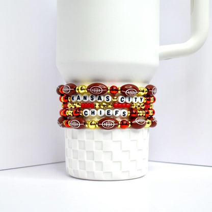 Stack of beaded bracelets wrapped around a white mug. The bracelets feature football-shaped beads, red and gold beads, and letter beads spelling 'KANSAS CITY CHIEFS'. The bracelets are red, gold, and brown with white lettering.