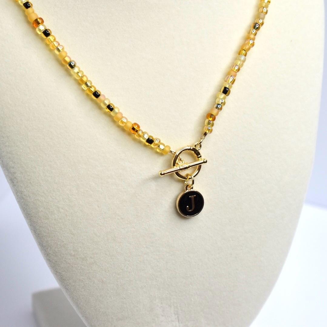 A beaded necklace with a gold-colored toggle clasp and a pendant. The necklace features small, round beads in shades of yellow, amber, and black. The pendant is a black circle with the letter 'J' in white. The necklace is displayed on a white surface.
