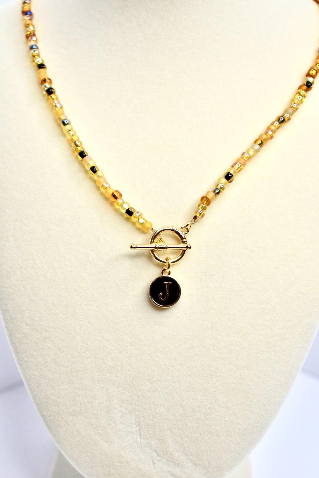 A gold beaded necklace with a toggle clasp and a circular pendant. The beads are a mix of yellow, gold, and black. The pendant is black with a gold border and the letter 'J' in gold. The necklace is displayed on a white surface.
