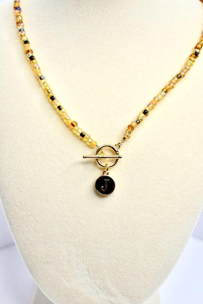 A gold beaded necklace with a toggle clasp and a circular pendant. The beads are a mix of yellow, gold, and black. The pendant is black with a gold border and the letter 'J' in gold. The necklace is displayed on a white surface.