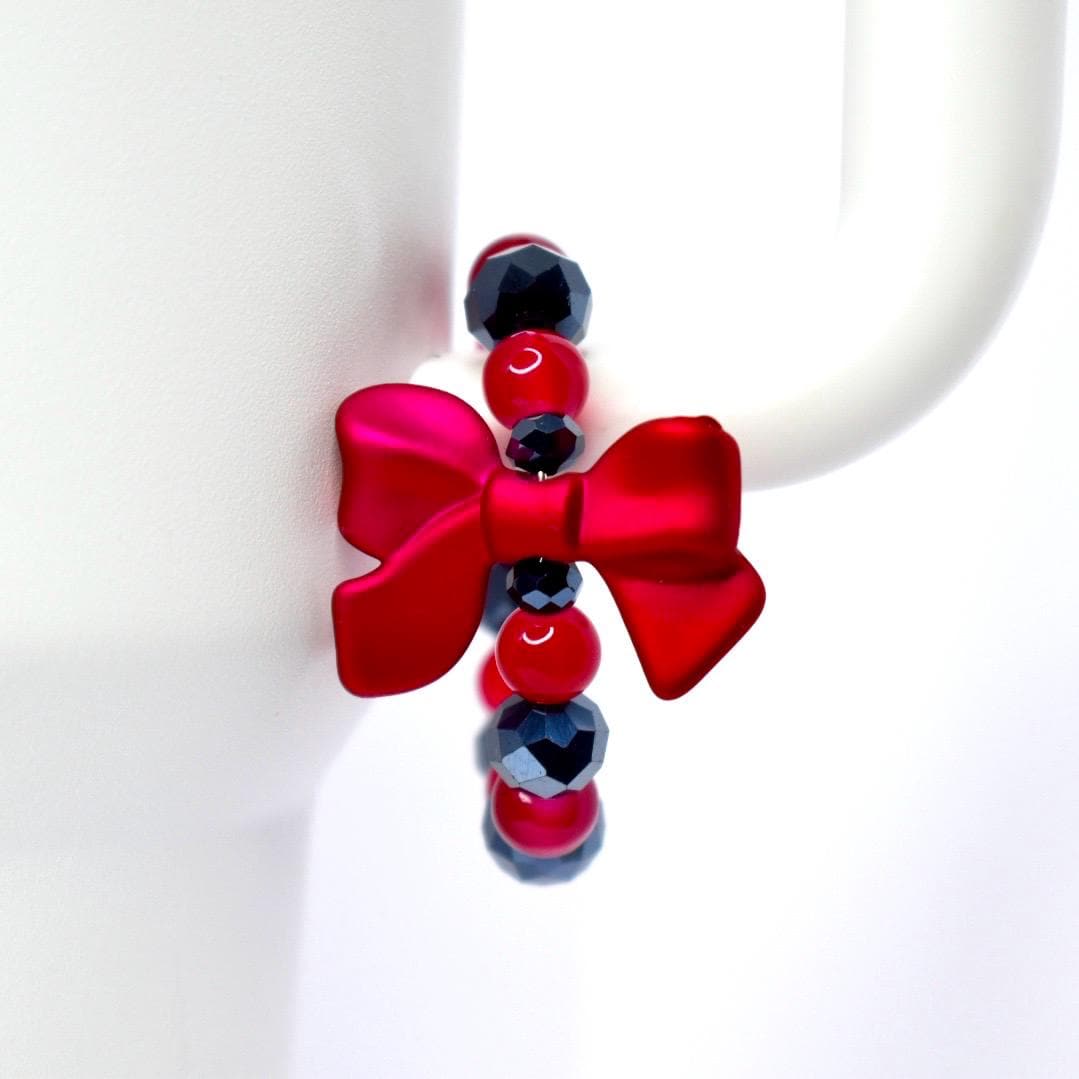 A red bow-shaped charm with a beaded strand of red and dark blue beads. The charm is attached to a white mug. The bow has a matte finish and the beads are round and faceted.
