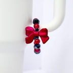 A red bow-shaped charm with a beaded bracelet. The bracelet features red, black, and dark blue beads. The charm is attached to a white mug handle. The bow is a vibrant, glossy red.