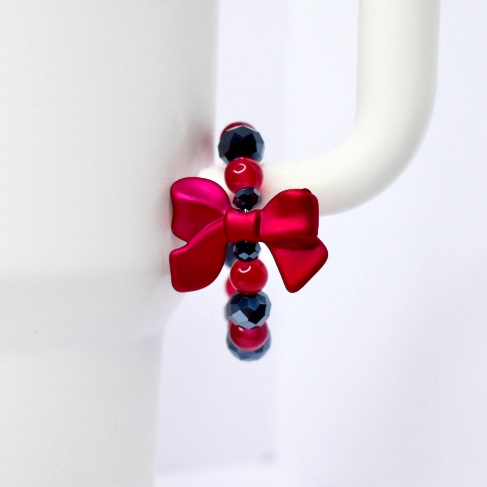 A red bow-shaped charm with a beaded bracelet. The bracelet features red, black, and dark blue beads. The charm is attached to a white mug handle. The bow is a vibrant, glossy red.
