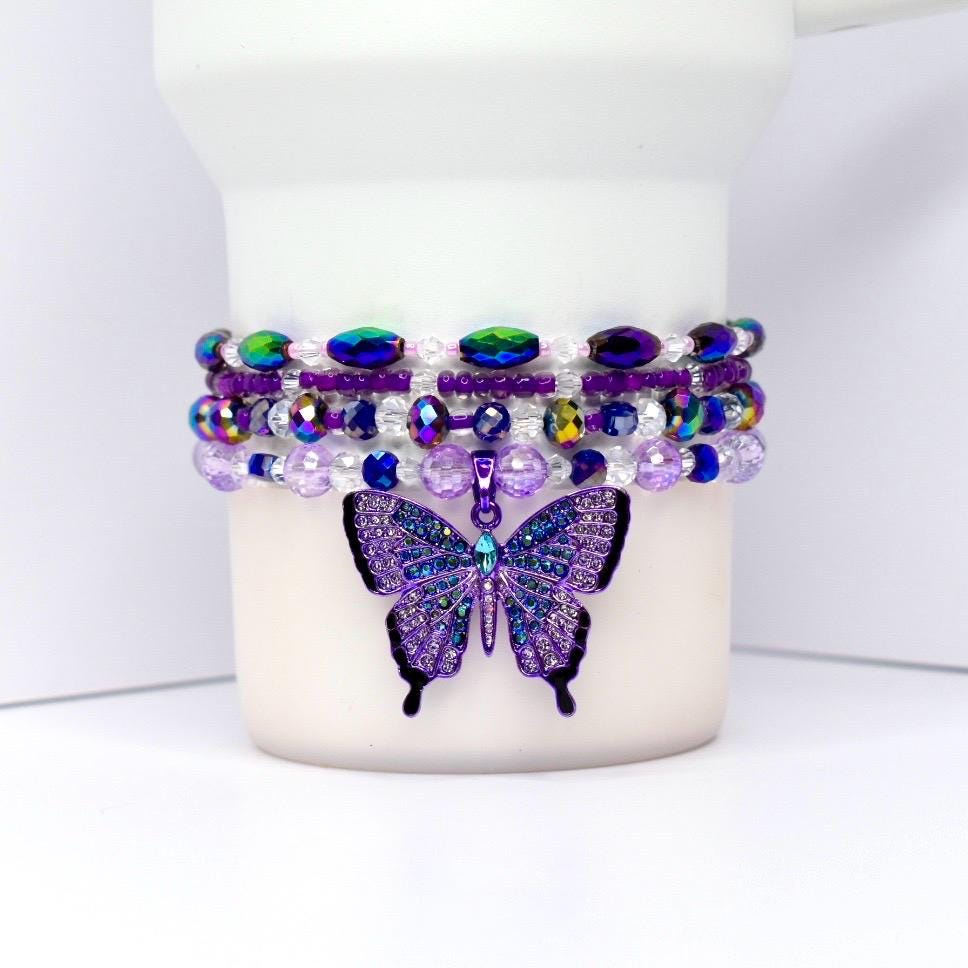 A stack of beaded bracelets in shades of purple, blue, and iridescent green, with a dangling butterfly charm. The butterfly charm is purple with blue and iridescent accents.