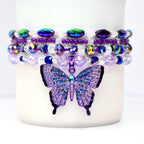 Four purple and iridescent stretch bead bracelets stacked around the base of a tumbler, featuring a rhinestone butterfly charm in the center.