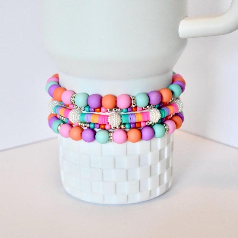 A stack of colorful beaded bracelets in shades of pink, orange, purple, and turquoise. The bracelets are adorned with small, sparkly accents and are displayed on a white, textured vase. The bracelets are a fun and vibrant accessory.