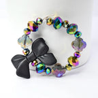 A bracelet with iridescent, faceted beads in shades of purple, green, and yellow. A black bow is attached to the bracelet, adding a contrasting element. The bracelet is displayed against a white background.