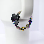 A black bow bracelet with iridescent beads. The bow is made of a matte black material. The beads are a mix of colors, including purple, gold, and blue, with a faceted design. The bracelet is displayed on a white surface.