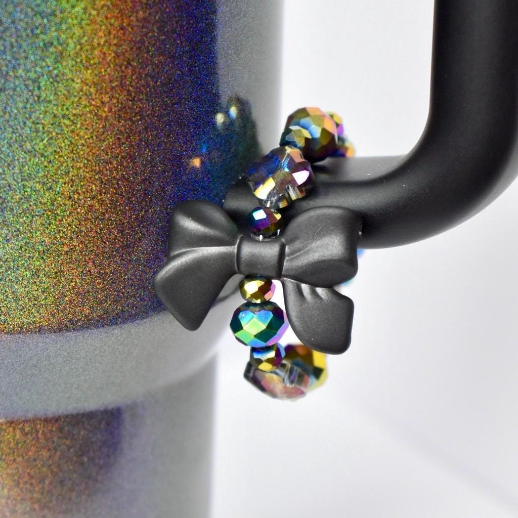A black bow-shaped accessory with iridescent beads attached to a black mug handle. The mug has a holographic, glittery finish with a rainbow of colors. The beads are a mix of colors, reflecting light. The bow is centered on the handle.