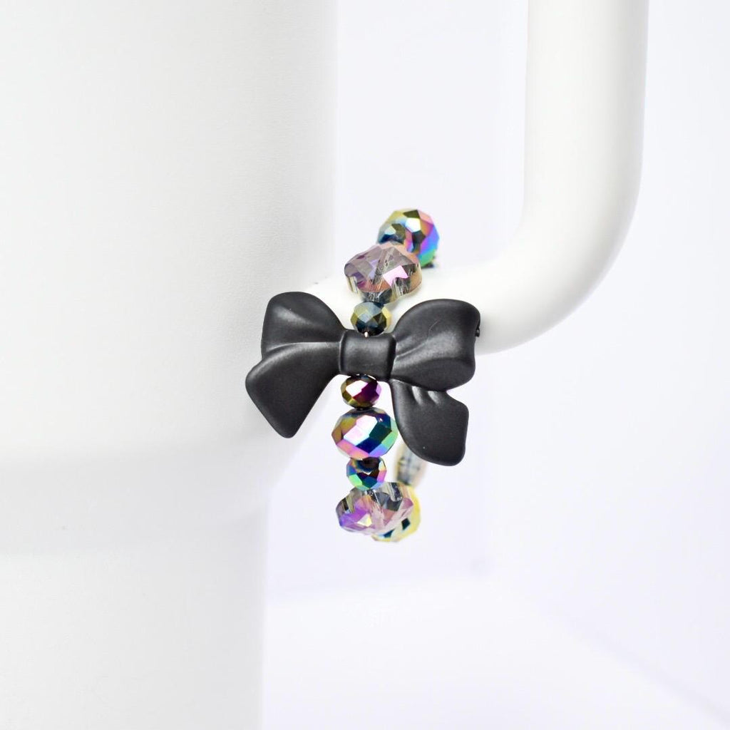 A black bow handle bracelet with iridescent beads. The bow is the focal point, made of matte black material. The beads are a mix of colors, reflecting light with a rainbow effect. The bracelet is displayed on a white Stanley, highlighting the contrast of colors and textures.