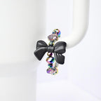 A black bow handle bracelet with iridescent beads. The bow is the focal point, made of matte black material. The beads are a mix of colors, reflecting light with a rainbow effect. The bracelet is displayed on a white Stanley, highlighting the contrast of colors and textures.