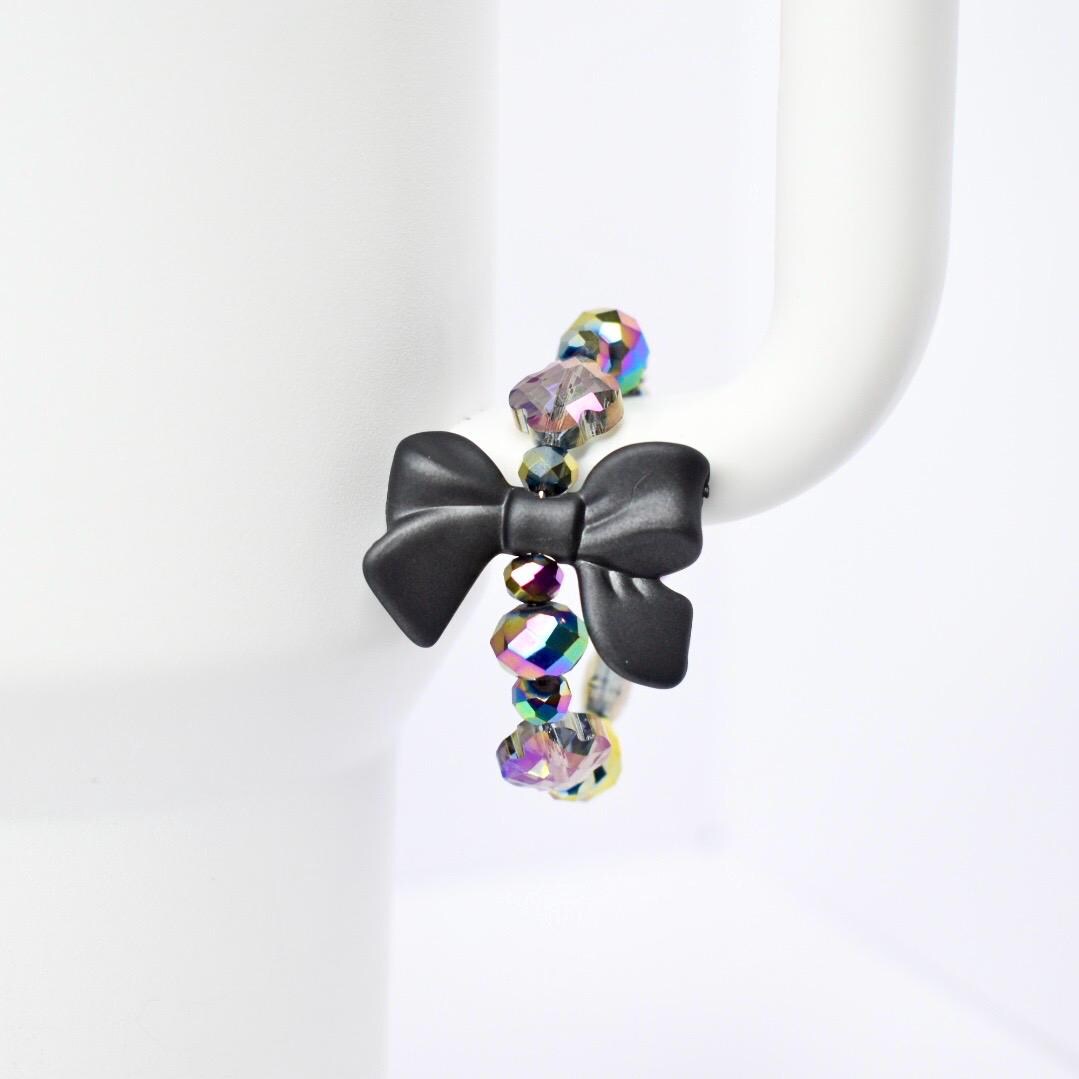 A black bow handle bracelet with iridescent beads. The bow is the focal point, made of matte black material. The beads are a mix of colors, reflecting light with a rainbow effect. The bracelet is displayed on a white Stanley, highlighting the contrast of colors and textures.