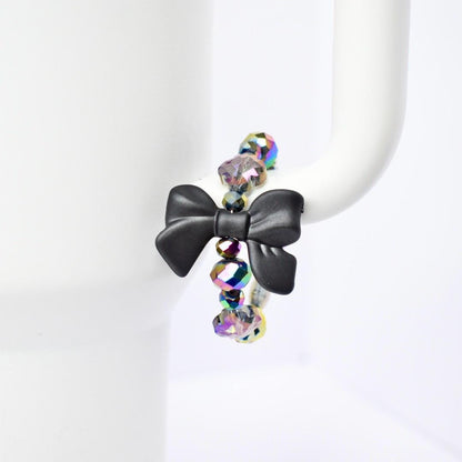 A black bow handle bracelet with iridescent beads. The bow is the focal point, made of matte black material. The beads are a mix of colors, reflecting light with a rainbow effect. The bracelet is displayed on a white Stanley, highlighting the contrast of colors and textures.