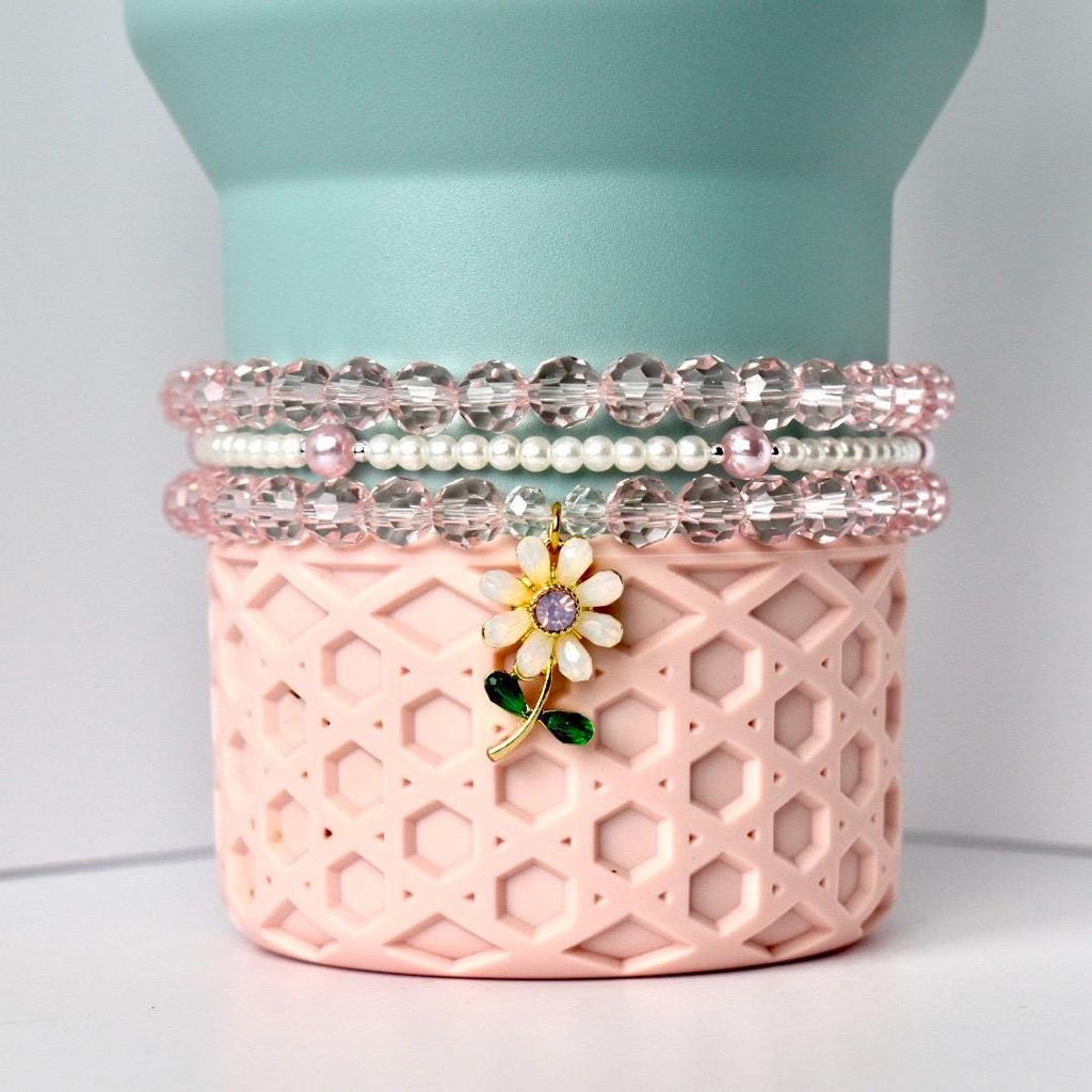 A stack of three beaded bracelets in shades of pink and clear. The bracelets are on a pink, patterned container with a flower charm. The charm has a white flower with a purple center and green leaves. The background is a light blue cylinder.