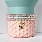 A stack of three beaded bracelets in shades of pink and clear. The bracelets are on a pink, patterned container with a flower charm. The charm has a white flower with a purple center and green leaves. The background is a light blue cylinder.