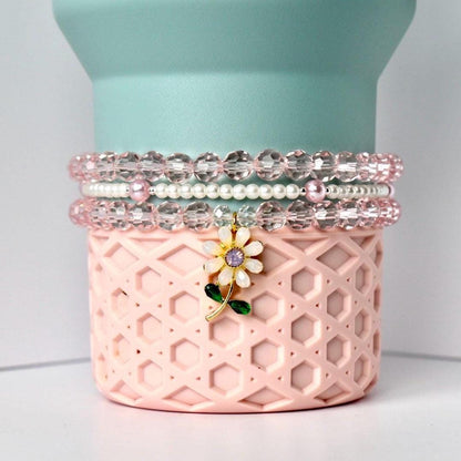 A stack of three beaded bracelets in shades of pink and clear. The bracelets are on a pink, patterned container with a flower charm. The charm has a white flower with a purple center and green leaves. The background is a light blue cylinder.