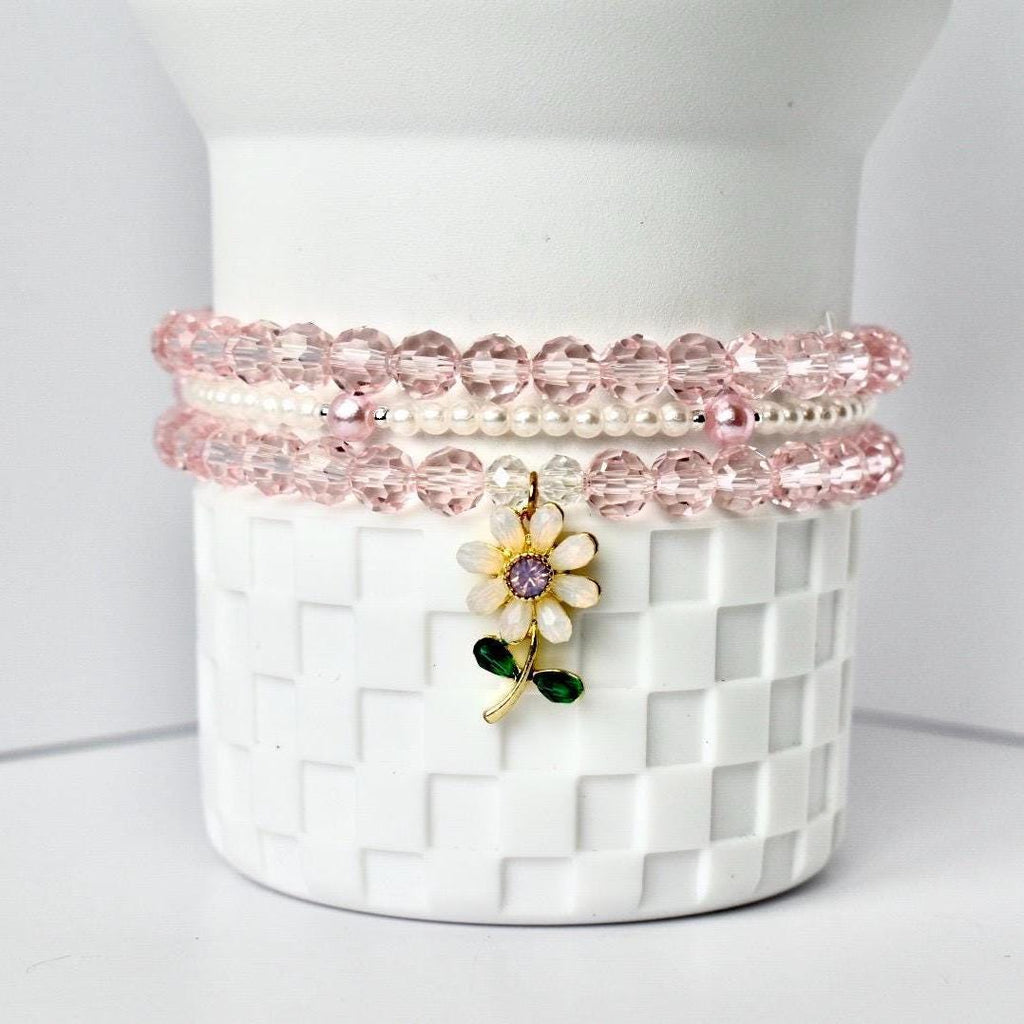 Three bracelets stacked on a white, checkered surface. The top and bottom bracelets are made of pink, faceted beads. The middle bracelet is made of small, white beads. A gold flower charm with a purple center and green leaves hangs from the bracelets.