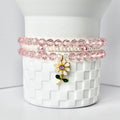 Three bracelets stacked on a white, checkered surface. The top and bottom bracelets are made of pink, faceted beads. The middle bracelet is made of small, white beads. A gold flower charm with a purple center and green leaves hangs from the bracelets.