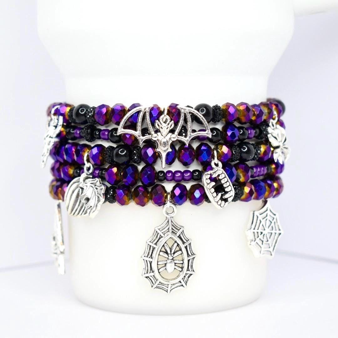 Stack of beaded bracelets in shades of purple, black, and gold. Each bracelet features a variety of charms, including a bat, spiderweb, and fangs. The bracelets are displayed on a white cylindrical object.