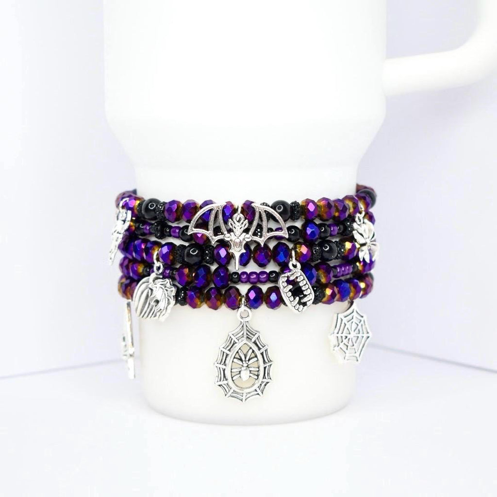 A stack of beaded bracelets in shades of purple, black, and iridescent colors. The bracelets feature silver charms including a bat, spiderweb, and fangs. The bracelets are displayed on a white mug.