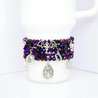 A stack of beaded bracelets in shades of purple, black, and iridescent colors. The bracelets feature silver charms including a bat, spiderweb, and fangs. The bracelets are displayed on a white mug.