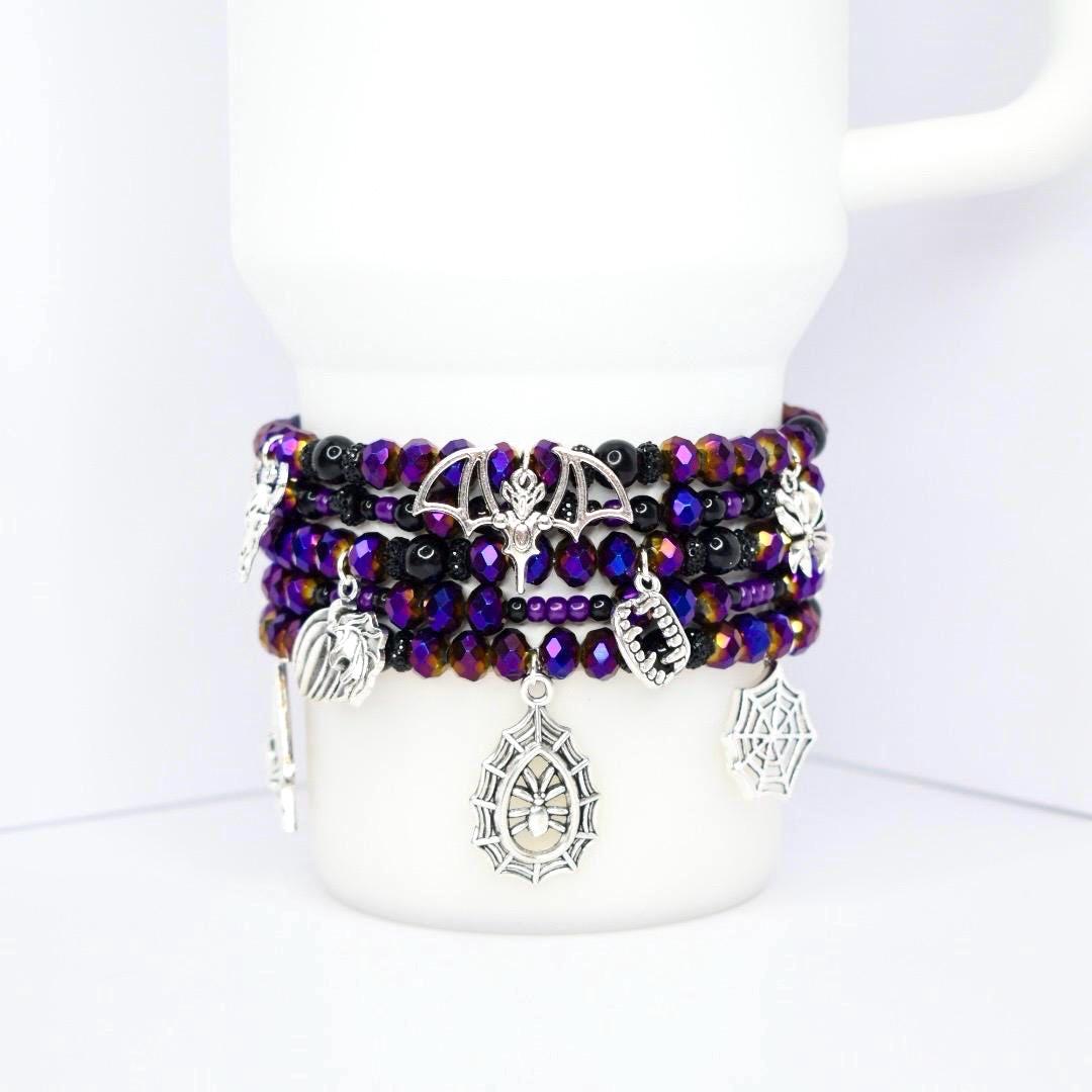 A stack of beaded bracelets in shades of purple, black, and iridescent colors. The bracelets feature silver charms including a bat, spiderweb, and fangs. The bracelets are displayed on a white mug.