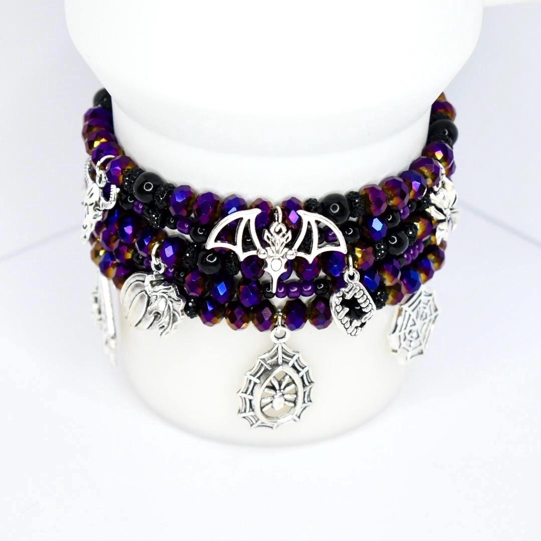 A stack of beaded bracelets with a gothic theme. The bracelets feature purple and black beads, with silver charms of bats, spiders, and spiderwebs. The bracelets are displayed on a white surface.