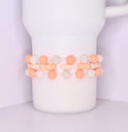 A set of three coordinated tumbler bracelets wrapped around a white tumbler, designed with soft peach matte beads, translucent crystal-like beads, textured white pearl beads, and flat cream-colored disc spacers.