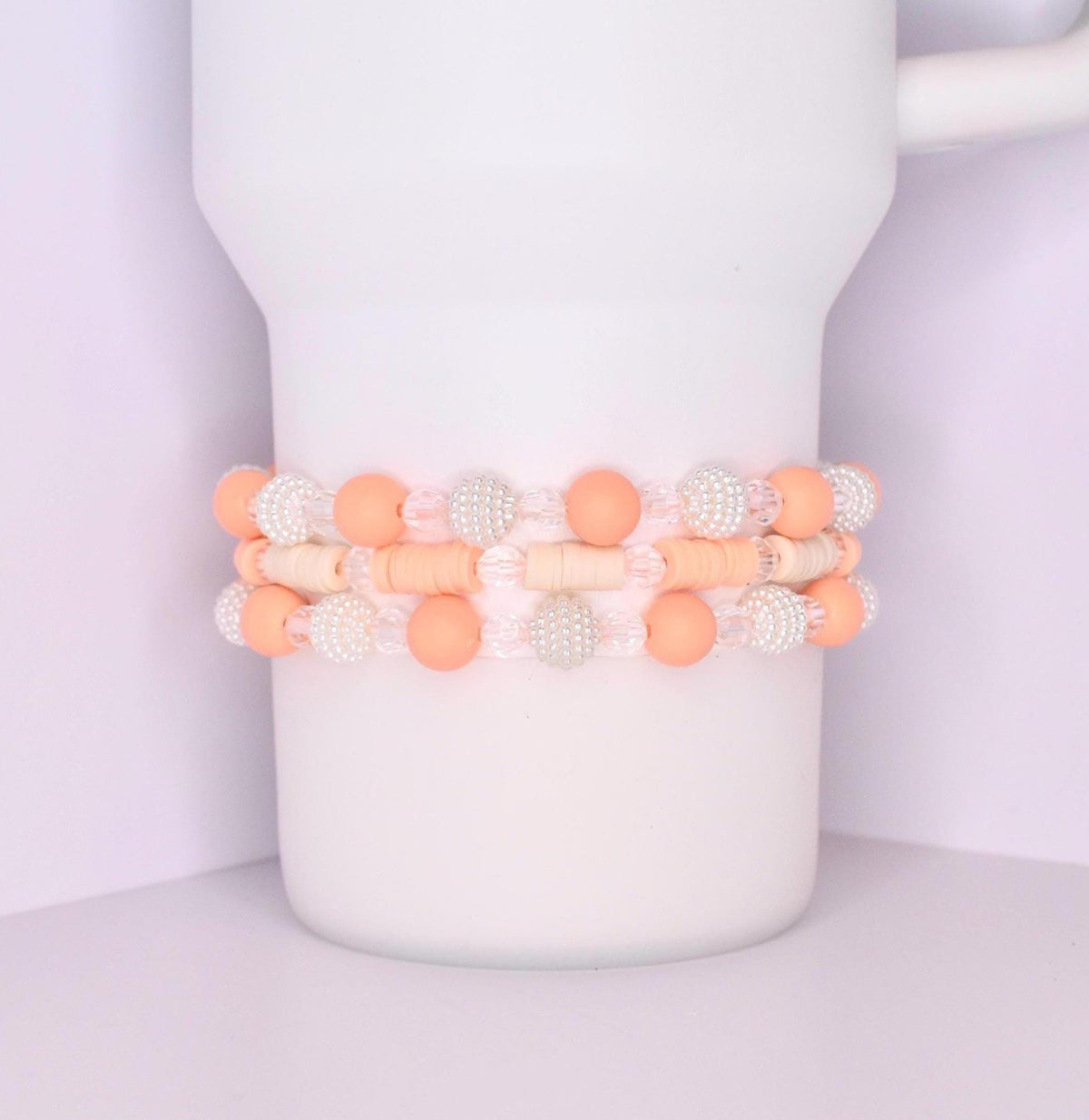 A set of three coordinated tumbler bracelets wrapped around a white tumbler, designed with soft peach matte beads, translucent crystal-like beads, textured white pearl beads, and flat cream-colored disc spacers.