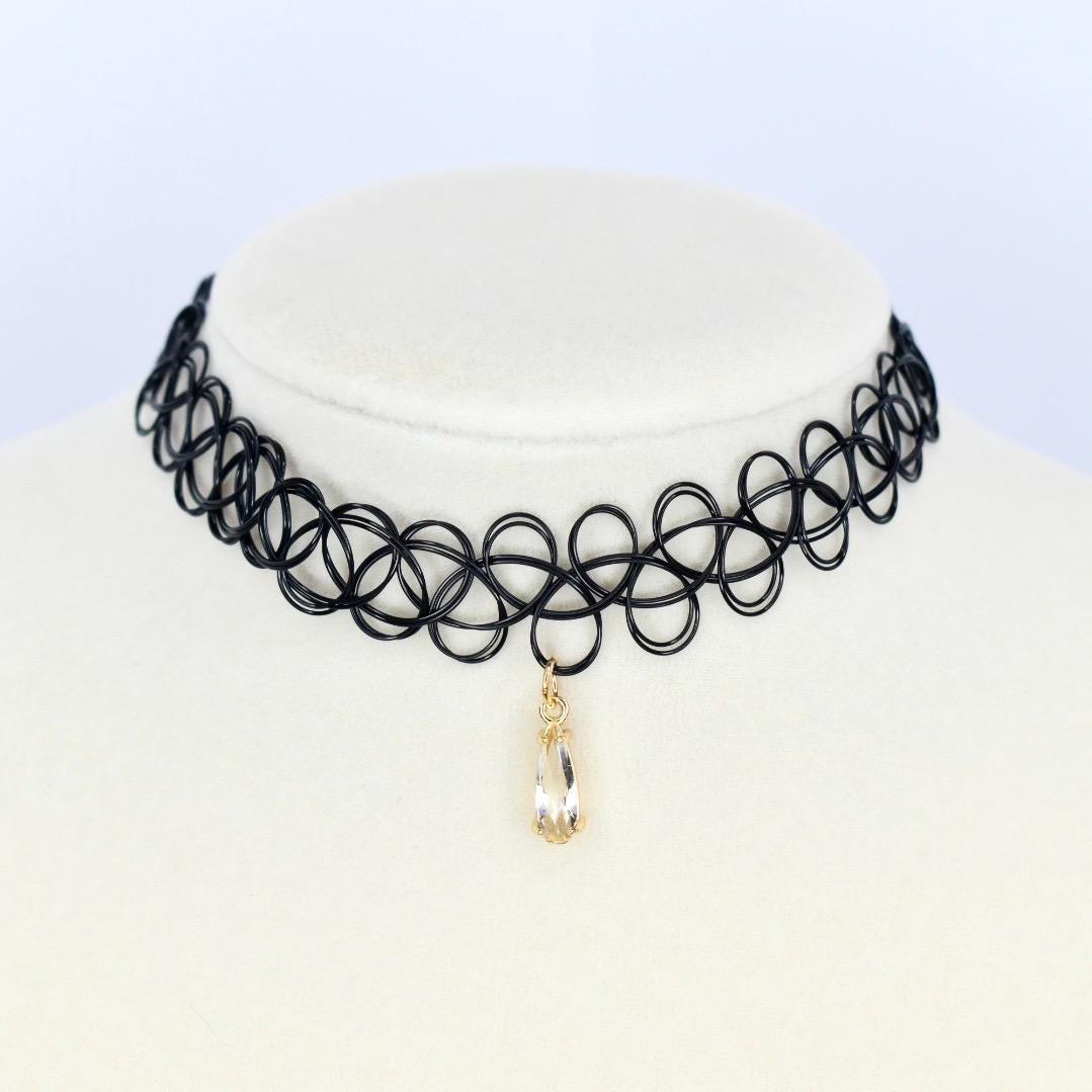 A black choker necklace with a pendant. The necklace is made of a series of interconnected black circles. A teardrop-shaped, clear crystal pendant hangs from the center of the choker. The pendant is encased in a gold-colored metal setting.